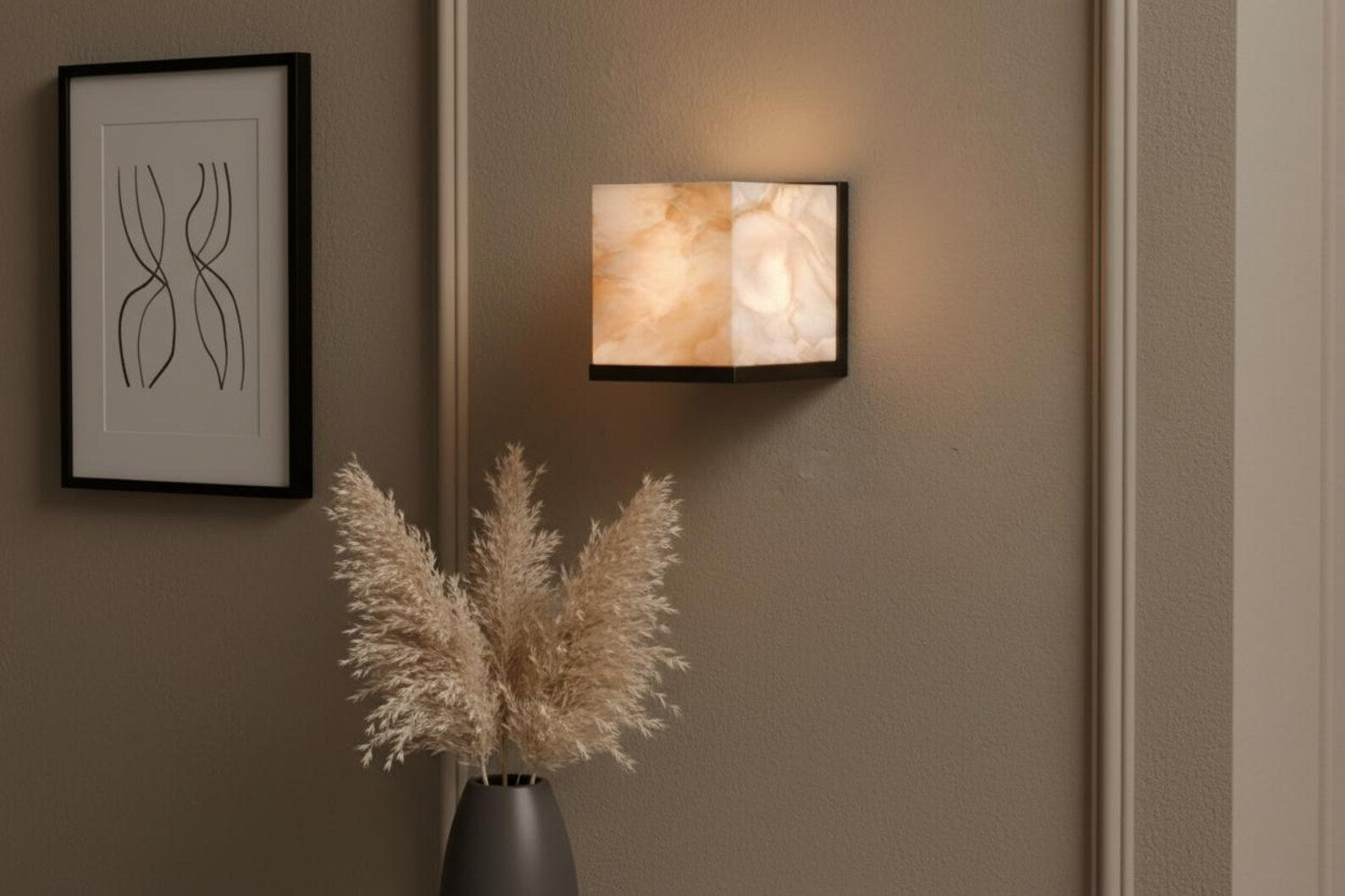 Wall-mounted light fixture on a wall with a vase of dried plants and a framed picture.