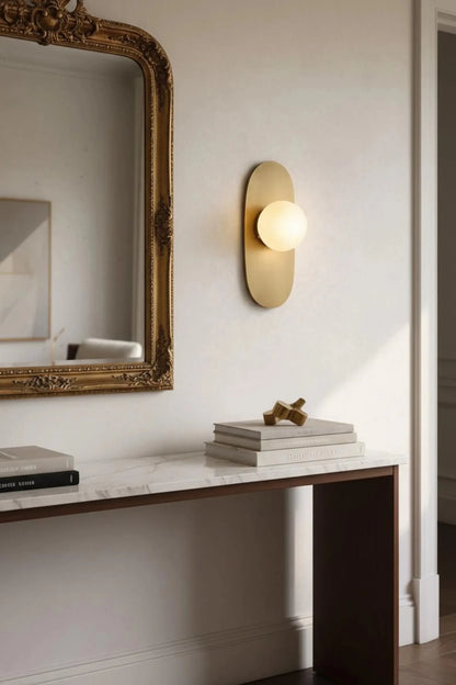 Decorative wall mirror and light fixture on a white wall with books on a surface.