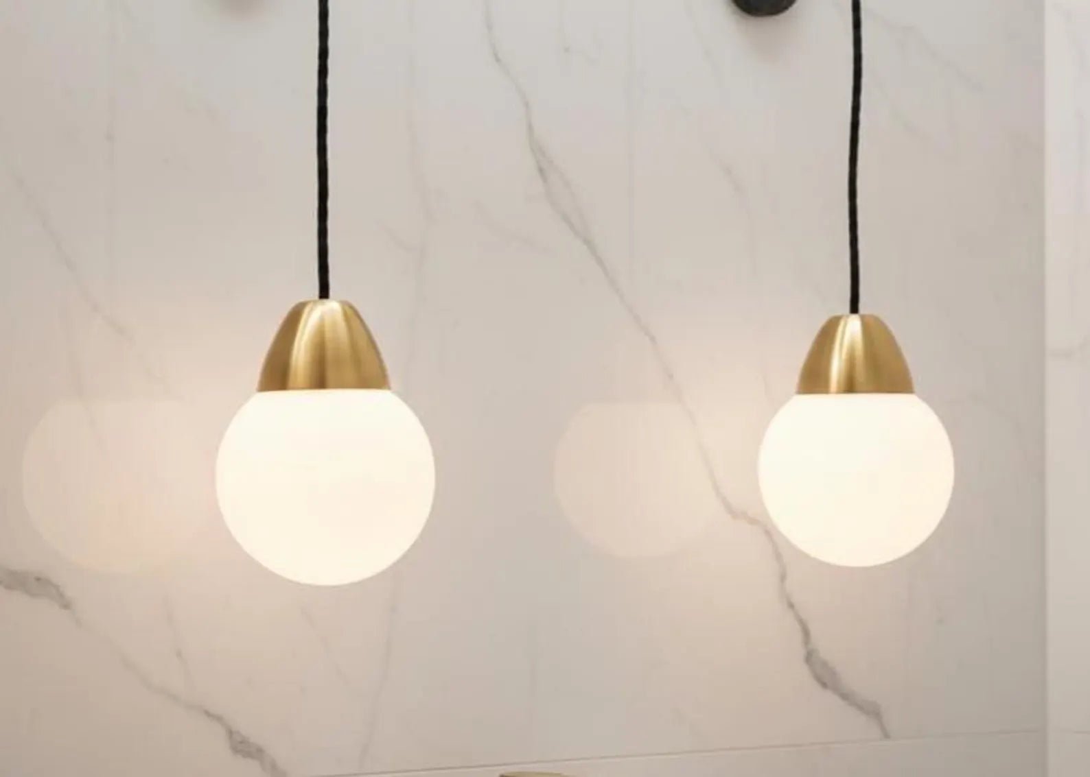 Two pendant lights with gold fixtures and white glass shades on a marble wall background.