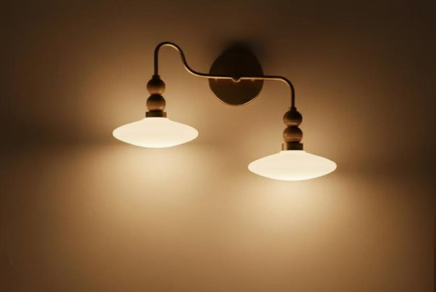 Wall-mounted lamp with two illuminated lampshades on a beige wall.