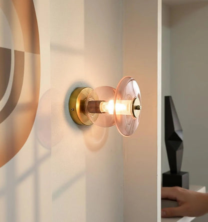 Wall-mounted light fixture with gold finish and clear glass shade on a neutral wall.