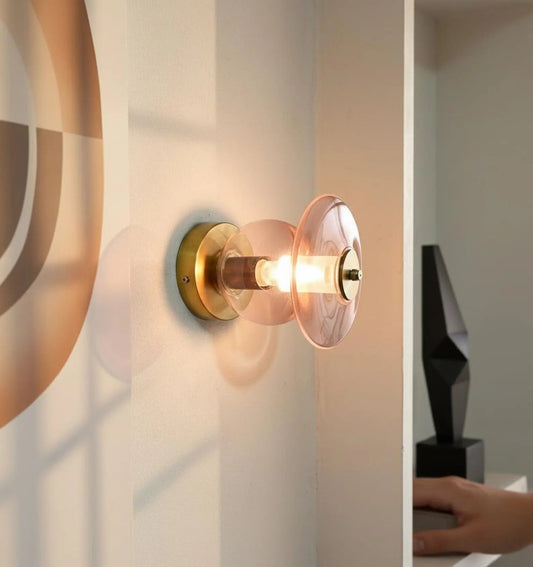 Wall-mounted light fixture with gold finish and clear glass shade on a neutral wall.