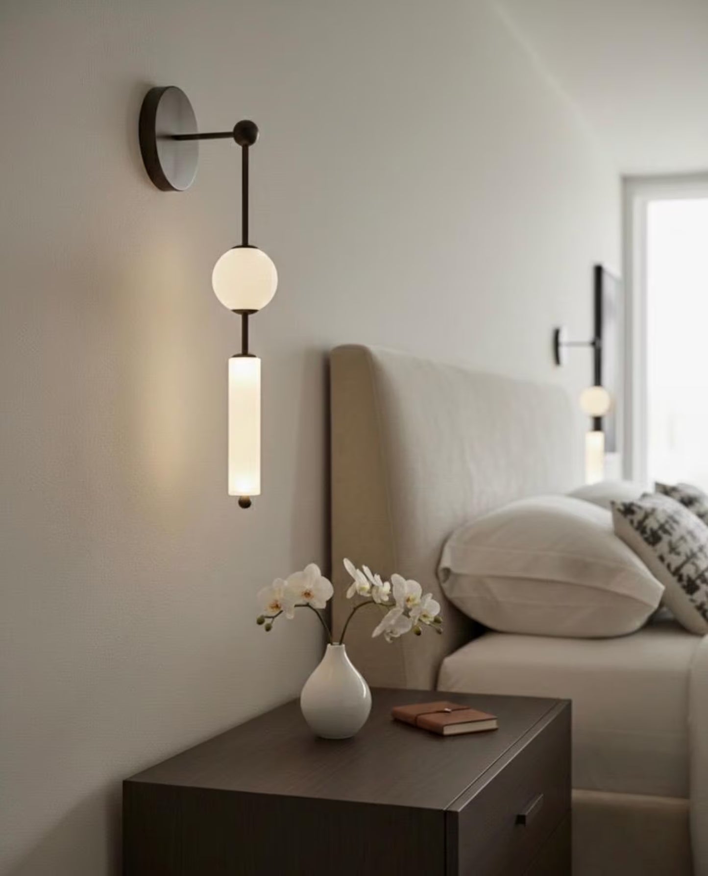 Modern wall sconce on a neutral wall with a vase of flowers and a book on a nightstand.